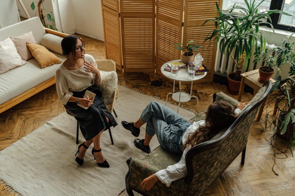 Zwei Frauen nehmen an einer Psychotherapiesitzung in einem warmen, einladenden Interieur mit Pflanzen und natürlichem Licht teil.