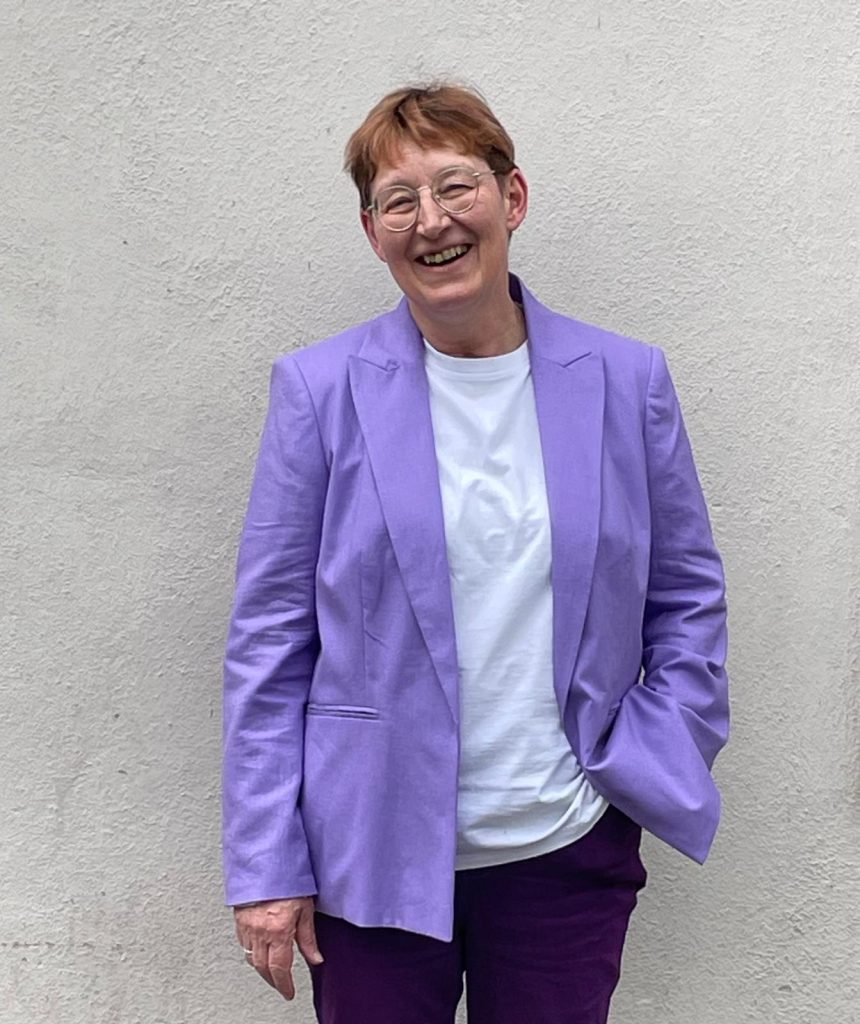 Eine Frau in einem lila Blazer und einer dazu passenden lila Hose steht selbstbewusst da und präsentiert ihr stylisches Outfit.